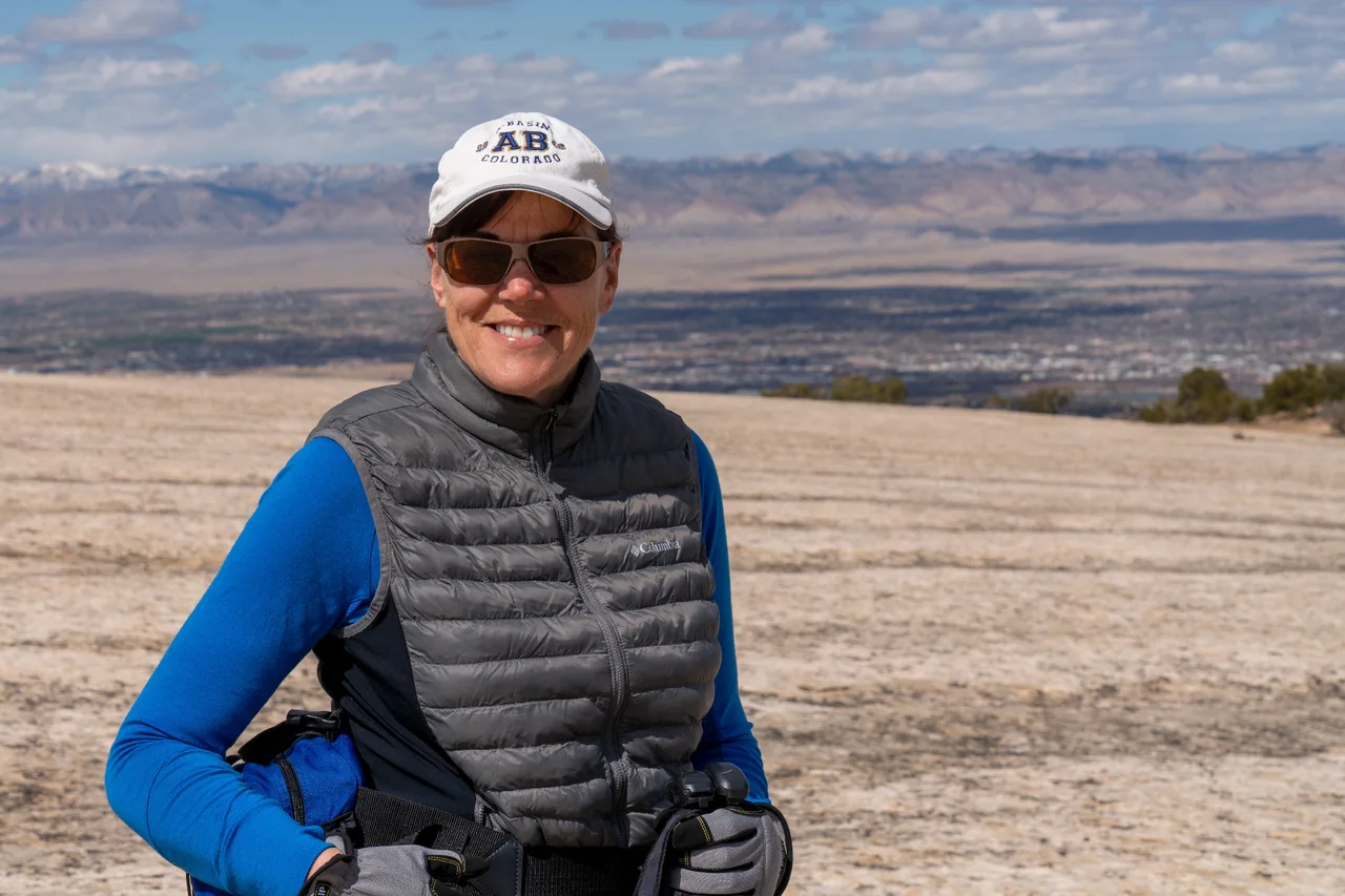 Adrienne smiling outdoors in sunglasses and a white cap, with a wide desert landscape behind her.