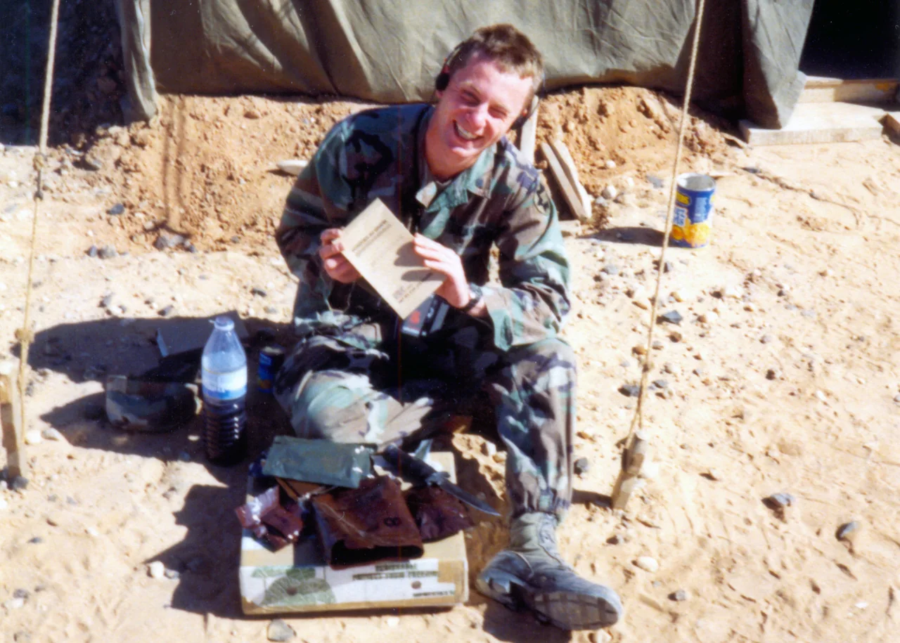 A younger Marcin in military uniform, seated outdoors in the desert and smiling with a book in hand.